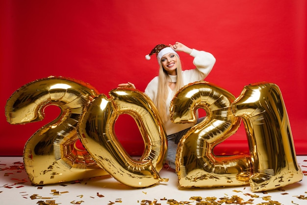 Xmas and New year celebrating young smiling girl in santa hat with golden balloons in 2021 shape on red background. High quality photo