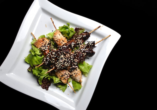 Yakitori with sesame seeds on a plate. Fried chicken meat and chicken liver on sticks close-up isolated on a black background. Top view.