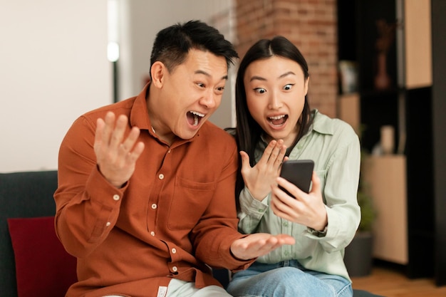 Yeah Emotional asian couple celebrating online win or great deal looking at smartphone with excitement