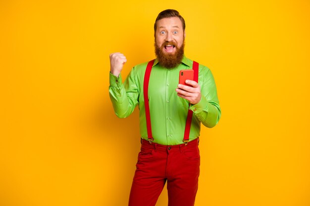 Yeah unbelievable! Crazy red hair redhair man use cellphone win social media lottery raise fists scream wear suspenders pants trousers isolated bright yellow color