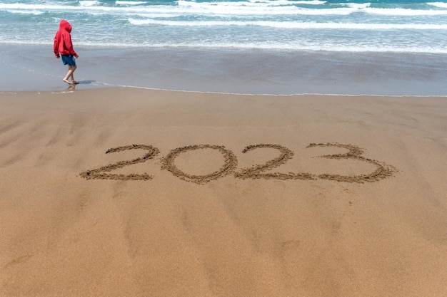 Year 2023 written in the sand on the beach with a boy in a red coat and the sea in the background