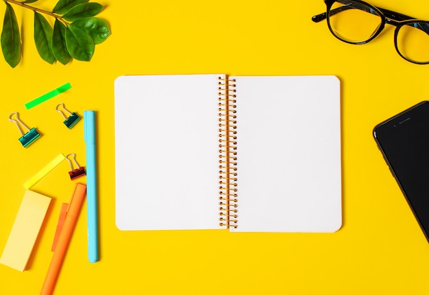 Yellow background, white notebook for records, phone, computer glasses, twig plants, pens pencils.