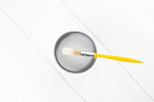 Yellow brush on an open can with white paint on a white wooden background top view