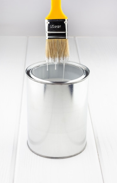 Yellow brush with white paint flowing into a can on a white wooden background closeup