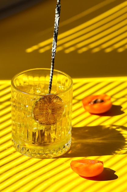 Yellow cocktail with ice in a clear glass with a bartender's spoon, lime chips and fresh apricot.