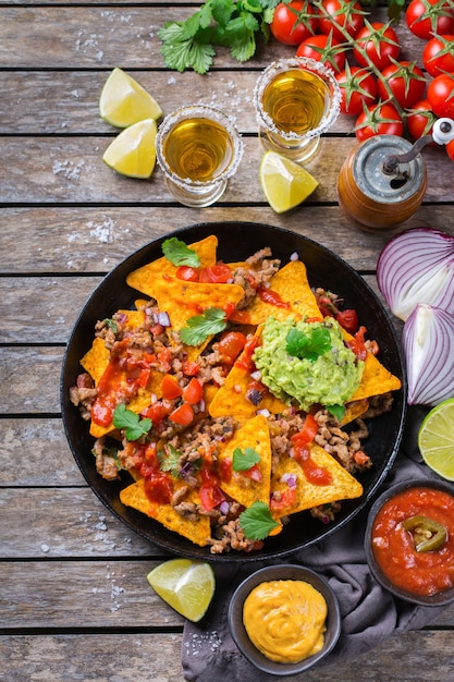 Yellow corn tortilla chips nachos with ground beef, mince, guacamole, red hot jalapeno chili salsa and cheese sauce with tequila on a wooden table. Top view flat lay background