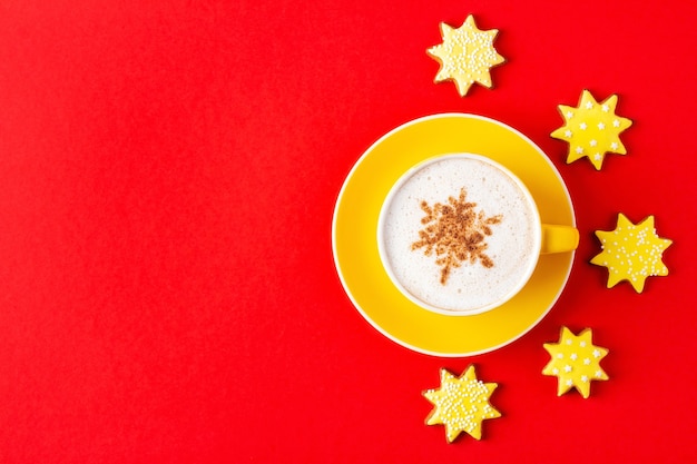 Yellow cup of Christmas Cappuccino on saucer and star shaped cookies on red background, flat lay. Holiday coffee with sweet treat.