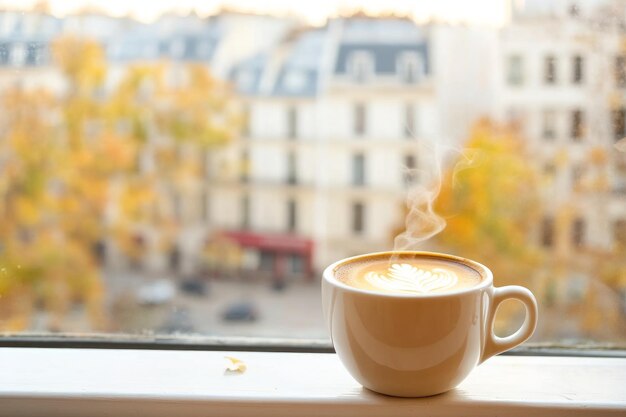 Photo a yellow cup of coffee with a soft steam on a windowsill in a bright autumn day blurred buildings of