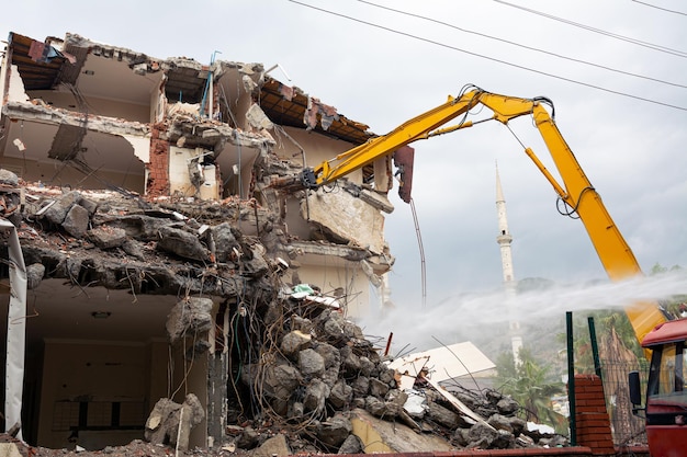 A yellow excavator is working on demolishing an old house in the city of Alanya Turkey A man in a truck with a barrel of water is pouring water on the rubble to keep it dustfree