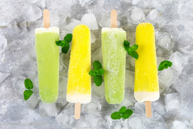 Yellow and green fruit ice cream on a stick with mint on ice on a light table.