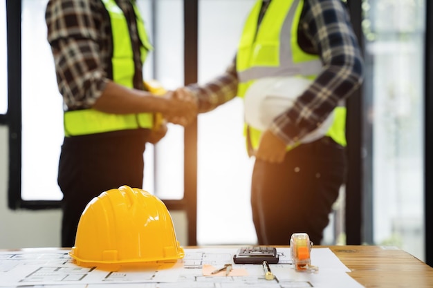 Yellow hard hat on workbench with construction worker hands shaking greeting start plan contracting new project in construction office center and contractor concept