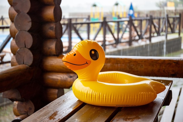 A yellow inflatable duck lies on massive wooden table in cozy gazebo of one of houses at recreation center against the backdrop of a large pool covered with heavy pouring rain