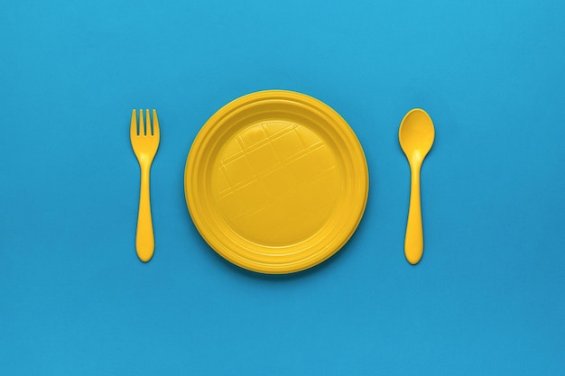 Yellow plastic plate and yellow spoon and fork on a blue background Minimal concept of plastic tableware Flat lay