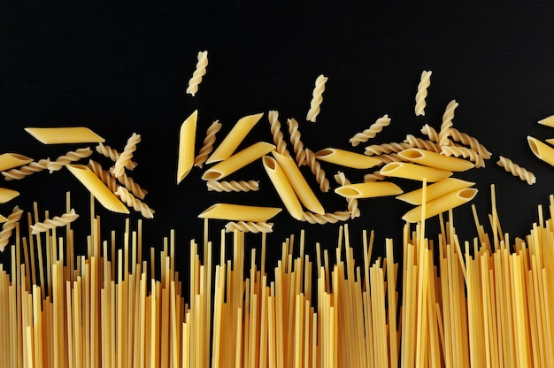 Yellow raw homemade spaghetti and penne pasta on a black concrete background at the bottom of the frame. Top view. The concept of cooking and diet.