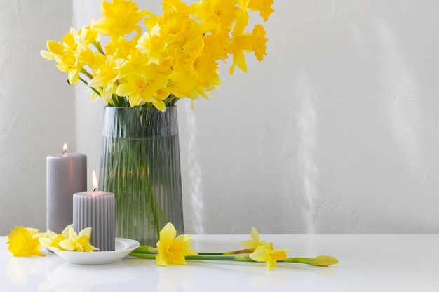 Yellow spring daffodils in glass vase with burning candles on white background