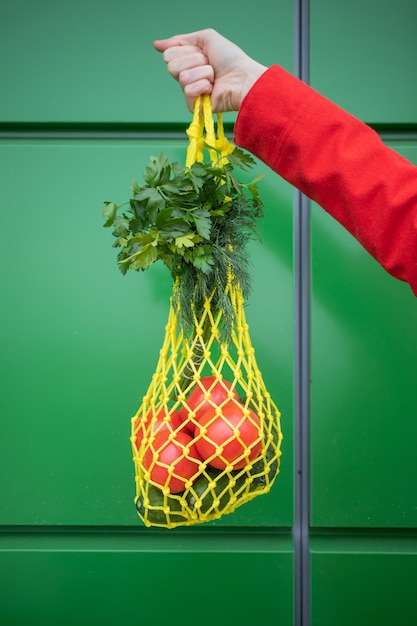 Yellow string bag with cucumbers, tomatoes, bananas and herbs in hand close-up. Bright photo in red, yellow, green tones. Sustainability, zero waste, plastic free concept, vegetarianism, healthy food.