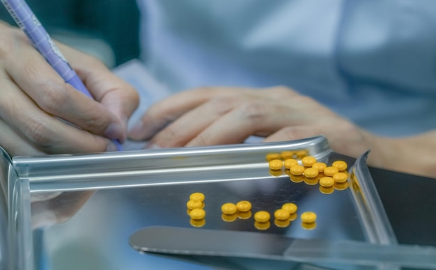 Yellow sugar coated tablet pill on stainless steel drug tray Pharmacist hand holding pen and writing