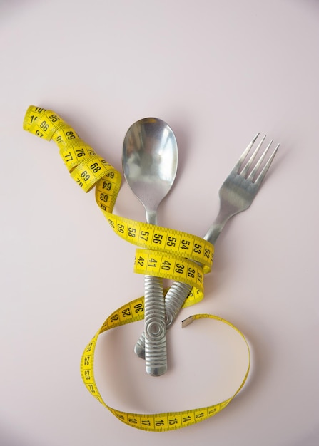 A yellow twisted centimeter ribbon is wrapped around a fork and spoon on a pink background