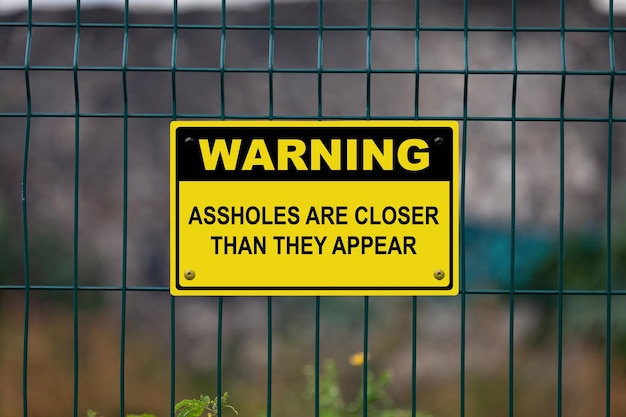 Photo yellow warning sign on a fence stating assholes are closer than they appear