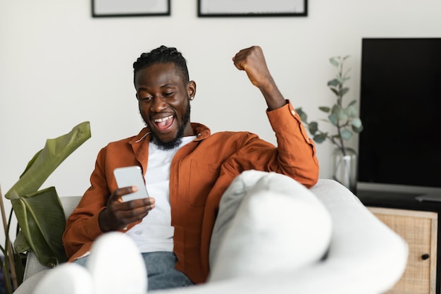 Yes great news Overjoyed african american man using smartphone and shaking clenched fist sitting on sofa