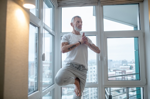 Yoga. Gray-haired man in a white tshirt standing in a tree pose