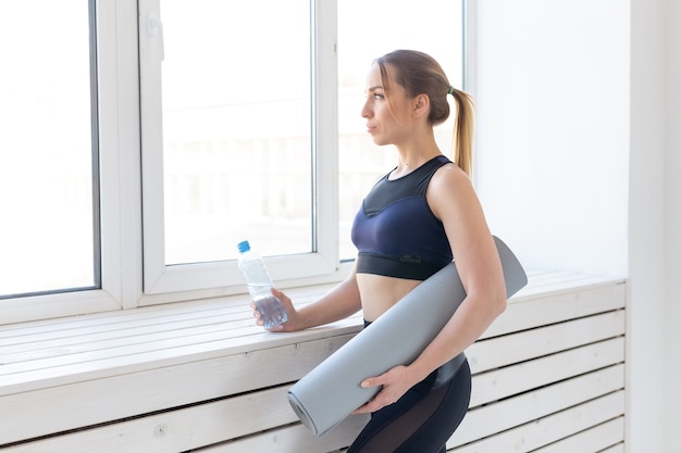 Yoga, healthy and sport concept - Young fit woman holding grey mat and bottle of water