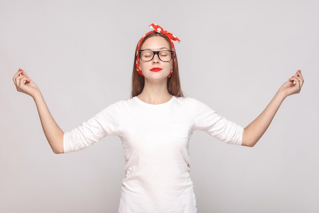 Yoga relaxed raised hands portrait of beautiful emotional young woman in white t-shirt with freckles, black glasses, red lips and head band. indoor studio shot, isolated on light gray background.