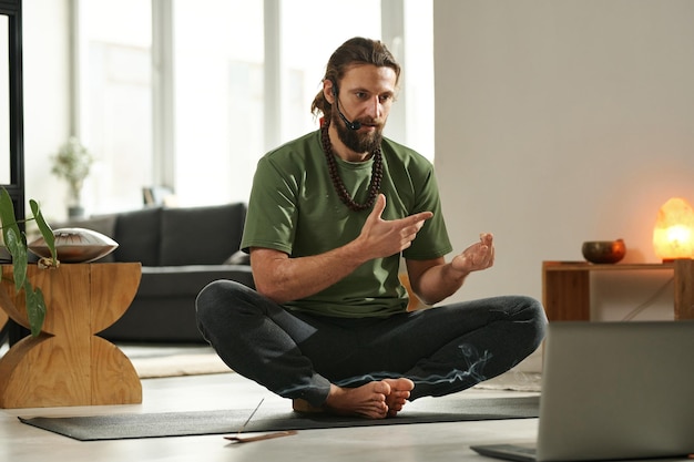 Yoga teacher sitting on exercise mat in front of laptop and having online communication with his stu