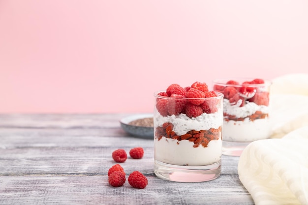 Yogurt with raspberry, goji berries and chia seeds in glass on gray and pink background and linen textile. Side view, copy space, selective focus.