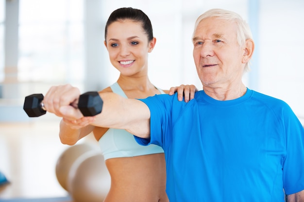 You are doing well! Cheerful female physical therapist helping senior man with fitness in health club