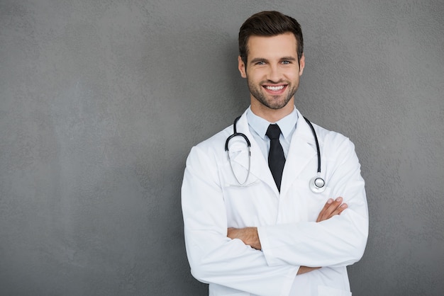 You can absolutely trust me. Confident young doctor in white uniform looking at camera and keeping arms crossed while standing against grey background