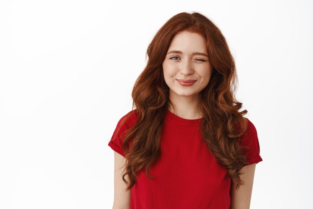 You got this Smiling redhead curly girl winking looking encouraging and supportive at camera standing in red tshirt against white background Copy space