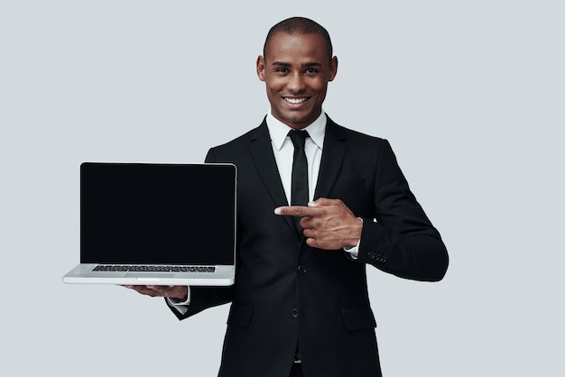 You should see this! Young African man in formalwear pointing at laptop and smiling while standing against grey background