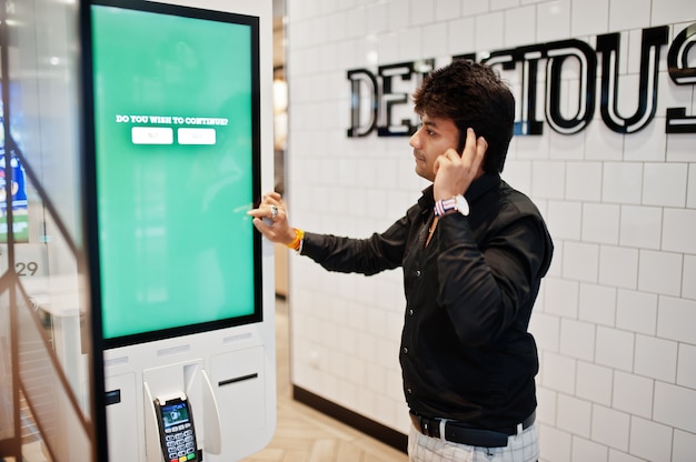 Do you wish to continue? Indian man customer at store place orders and pay through self pay floor kiosk for fast food, payment terminal. His thinking of choise.