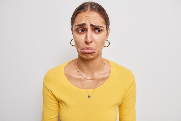 Youg brunette European woman feels very upset frowns eyebrows and purses lips looks lamentable feels frustrated wears casual yellow jumper isolated over white background. Negative emotions concept