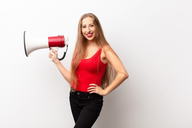 Yound blonde woman smiling happily with a hand on hip and confident, positive, proud and friendly attitude holding a megaphone