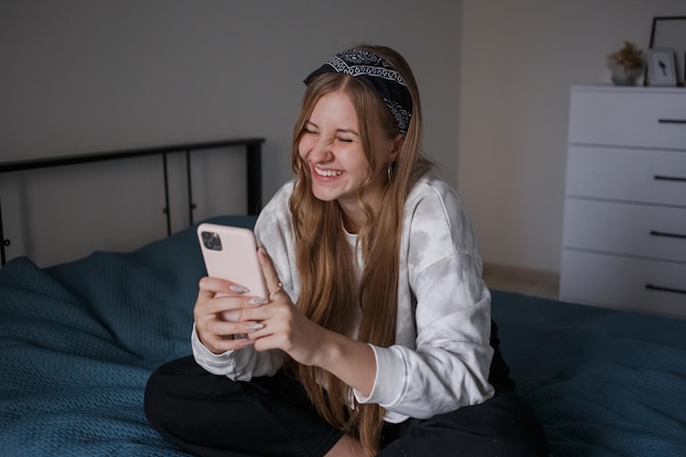 A young 20s girl  sits on a bed in her living room holds a phone in her hands and laughs