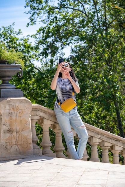 Young 30 years old woman in jeans tshirt and yellow bag walking in a park Ciutadella in Barcelona and taking photo on her smartphone Traveling in Spain