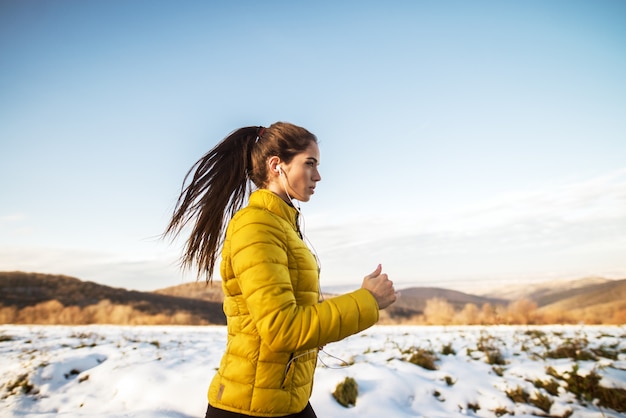 Young active athletic girl jogging in winter sportswear on snowy winter road with earphones in the sunny morning.