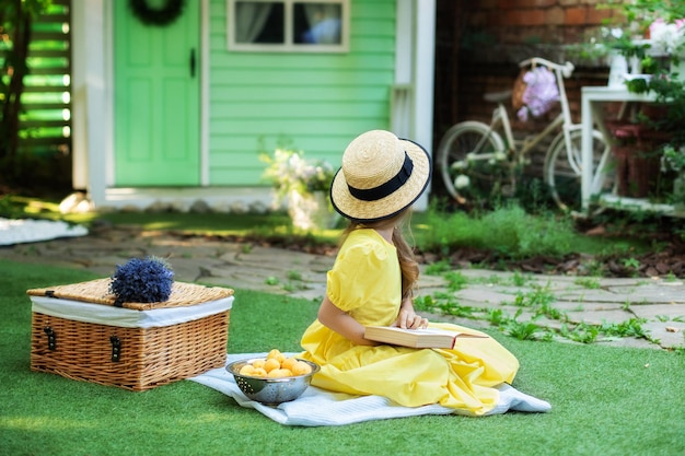 Young adorable girl in hat sitting on lawn in park with book in hands at summer.