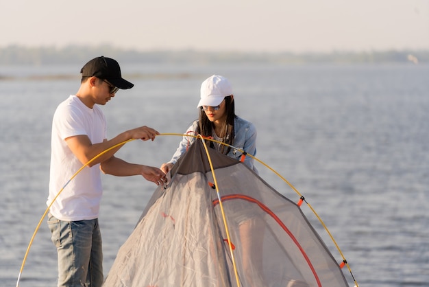 Young adult Asian couple pitch and set up tent for camping around the lake.