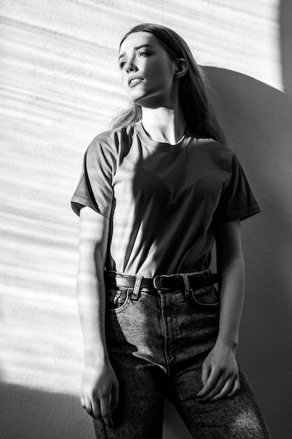 Young adult beautiful woman wearing Tshirt and jeans standing looking away with pensive romantic facial expression Black and white photography indoor studio shot illuminated by sunlight from window