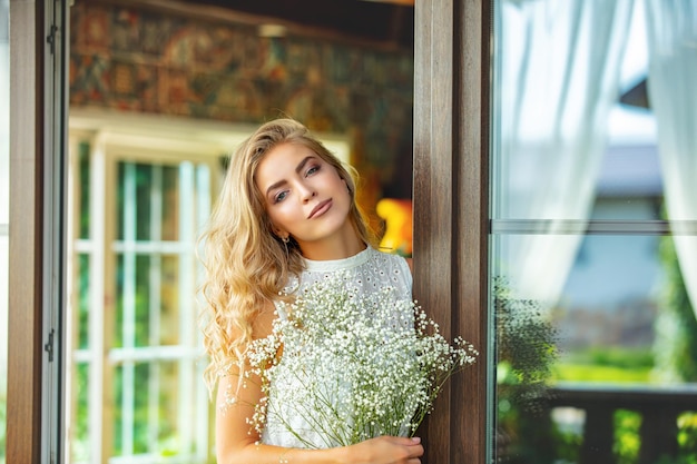 Young adult blonde beauty in a white dress at the windows on the balcony with white flowers