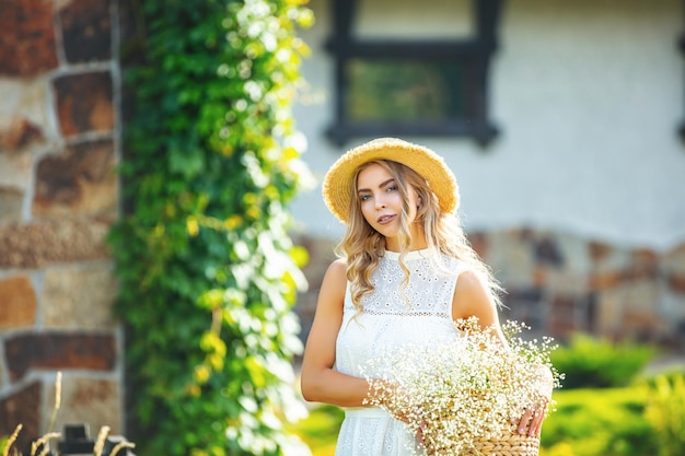 Young adult blonde in a white dress with a basket of flowers in the garden at house