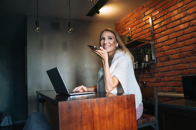 Young adult blonde woman with long hair working on laptop and using mobile phone on kitchen at home remote work