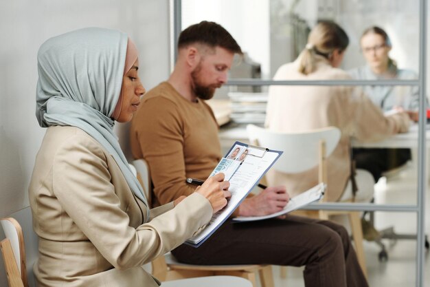 Young adult caucasian man and muslim woman in hijab sitting on chairs in visa service agency filling