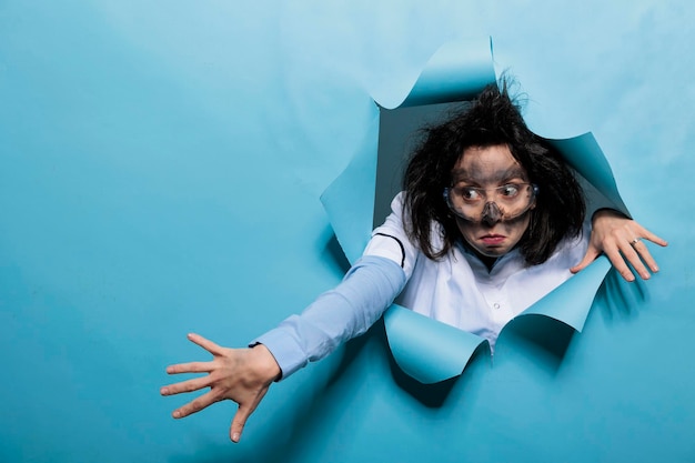 Young adult chemist with wacky look, messy hair and dirty face acting goofy after experiment explosion. Foolish humorous scientist with comic expression on blue background. Studio shot.