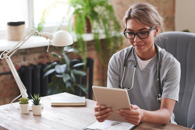 Young adult female doctor wearing eyeglasses sitting at desk holding digital tablet listening to pat