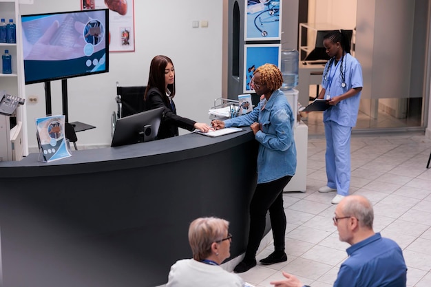 Young adult filling in registration form at hospital reception desk, writing medical checkup report before attending appointment. Woman checking papers in facility lobby, having consultation.