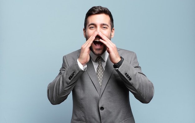 Young adult handsome businessman feeling happy, excited and positive, giving a big shout out with hands next to mouth, calling out